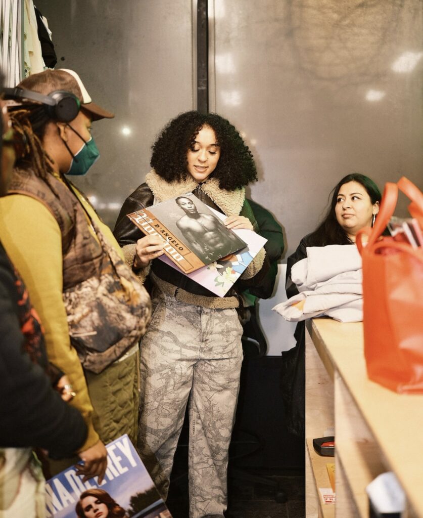 Several participants are gathered in the corner of a room, holding their records at the Vinyl Collectiv event Bring a Record, Catch a Vibe. One person is wearing a mask and headphones while holding a Lana Del Rey vinyl. Another person holds the D'Angelo vinyl, Voodoo. Photograph by Austin Scott.