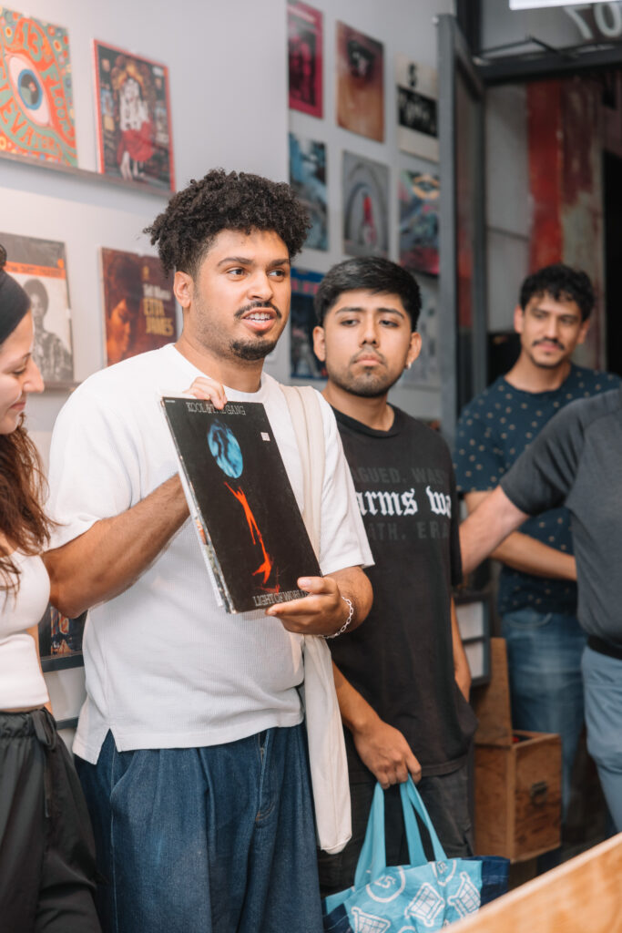 A person in a white t-shirt holds a record up to show a group of people at the Vinyl Collectiv event Bring a Record, Catch a Vibe. Records are displayed on the wall behind the group. Photo by Wendy Davis.