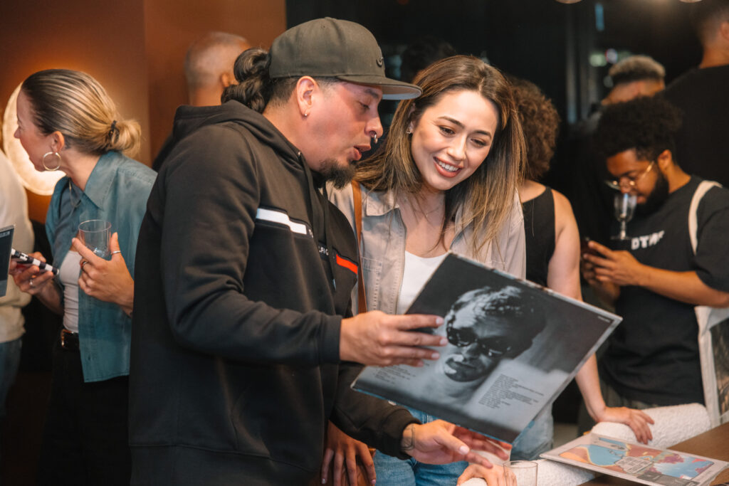 Two people talk while looking at a record together. Behind them is a crowd of people holding drinks and talking. Photo by Wendy Davis.