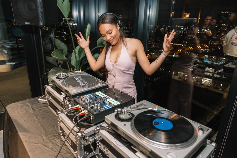Virginia stands at a DJ table with her hands thrown up in the air as she spins records at an event.  Photo by Wendy Davis.
