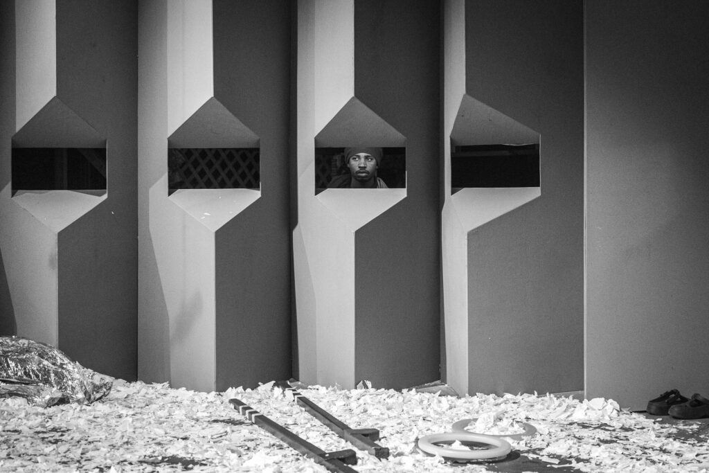 Theatre Y ensemble member Makai Walker in Charges (The Supplicants). A person is looking through a viewing window cut into a grey wall. The floor is covered in torn bits of paper and littered with shoes, stilts, and a reflective blanket. Image by Karl Soderstrom.