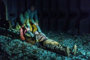 Two masked figures drag an unmasked person by their arms across a floor covered in torn bits of paper.