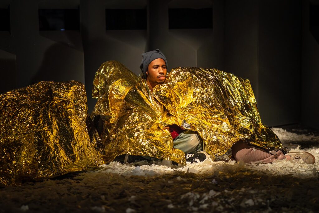 Theatre Y ensemble member Makai Walker and several other ensemble members in Charges (The Supplicants.) An unmasked person with a somber expression is sitting on a floor covered with torn bits of paper. They are surrounded by other figures who are asleep underneath reflective golden rescue blankets. Image by Karl Soderstrom.