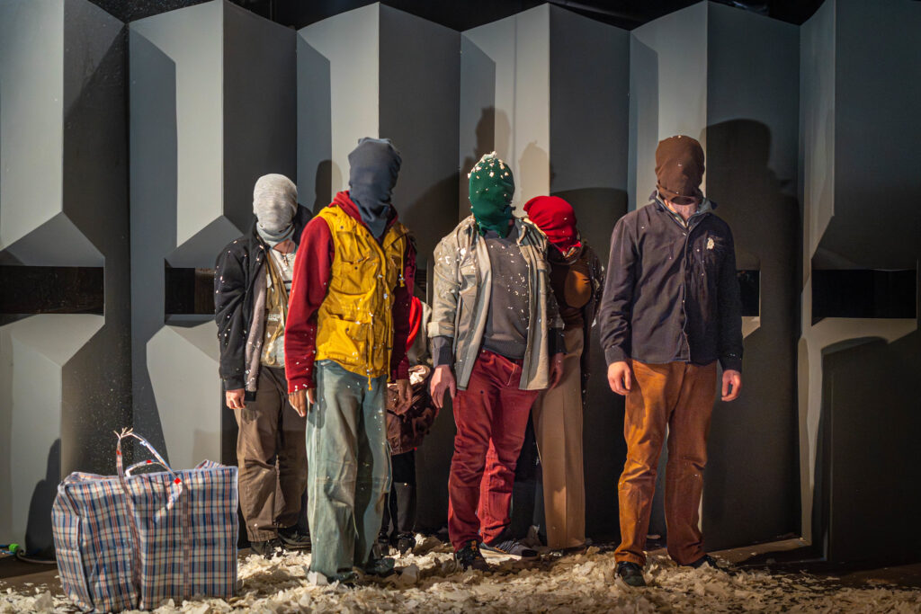 Five masked figures stand in front of a gray wall with viewing windows. The floor is covered with torn bits of paper and a large checkered bag is beside the figures. Photo by Karl Soderstrom.