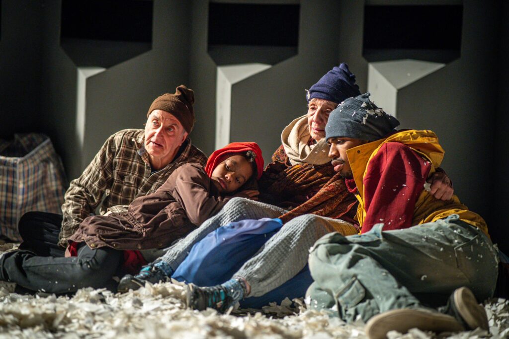 Theatre Y ensemble members Michael Awe, Kyndal Keith, Arlene Arnone, and Makai Walker in Charges (The Supplicants). Four people huddle together on a concrete floor covered with bits of torn paper. A gray wall with viewing windows is visible behind them. Image by Karl Soderstrom.