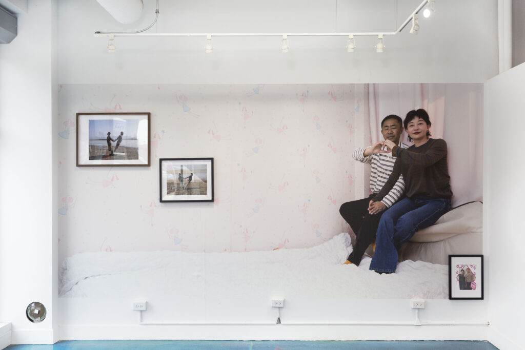 Installation view of Yangtze Love. Wallpaper image of a couple sitting next to each other in a pale, pink room. Together  they create a heart with their hands (each person only uses one hand for their half of the heart), while the other hands are joined together. In the upper, left corner are two framed images of people using their full bodies to make the shape of a heart. In the bottom right corner is another framed image of the couple, standing side by side surrounded by pink hearts. Photo courtesy of the artist.