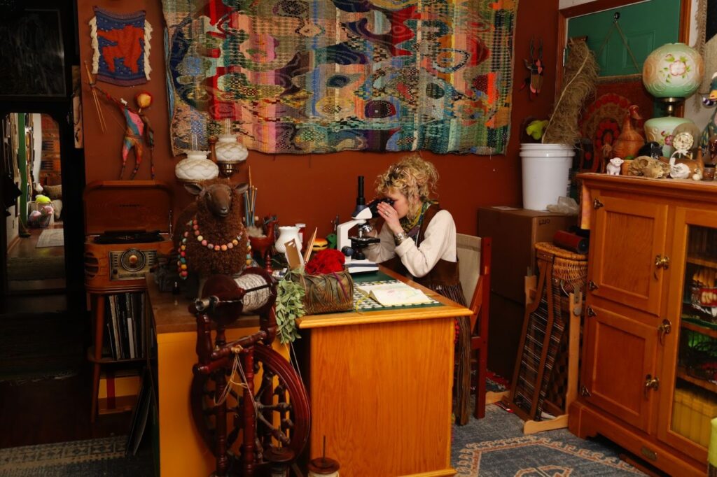 Rachel sits at her home desk and peeks through a microscope. “Pellicle Cartography”, a woven drawing of a pellicle, is hanging in the background and features nearly every color imaginable in an array of static-meets-form. Small, homey trinkets are seen throughout the space, as well as lots of warm-toned wood, a spinning wheel, and a record player. Image by Viki Stark.