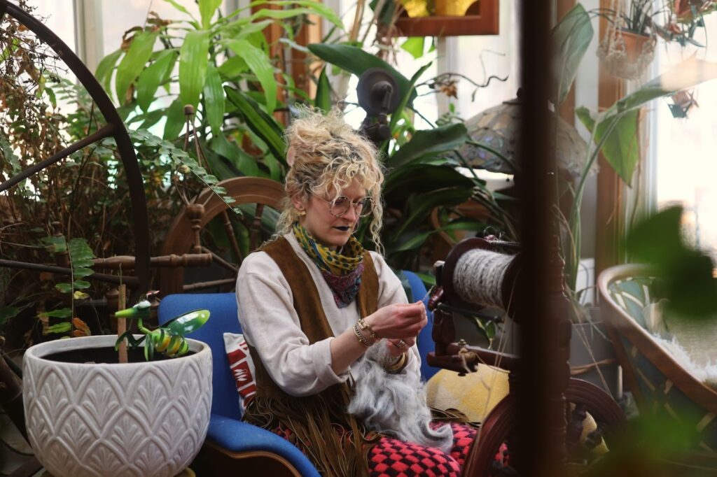 Rachel sits with her hair up in a clawclip while spinning yarn on the wheel in her house. She wears a brown fringed vest and her grandma’s colorful silk scarf around her neck. Many potted plants fill the room, their green leaves leaning throughout the background. The raw wool on Rachel’s lap is from Fiber Fleet and is black Icelandic wool combed together with white Mohair from her goat, Cammie. Image by Victoria Stark.