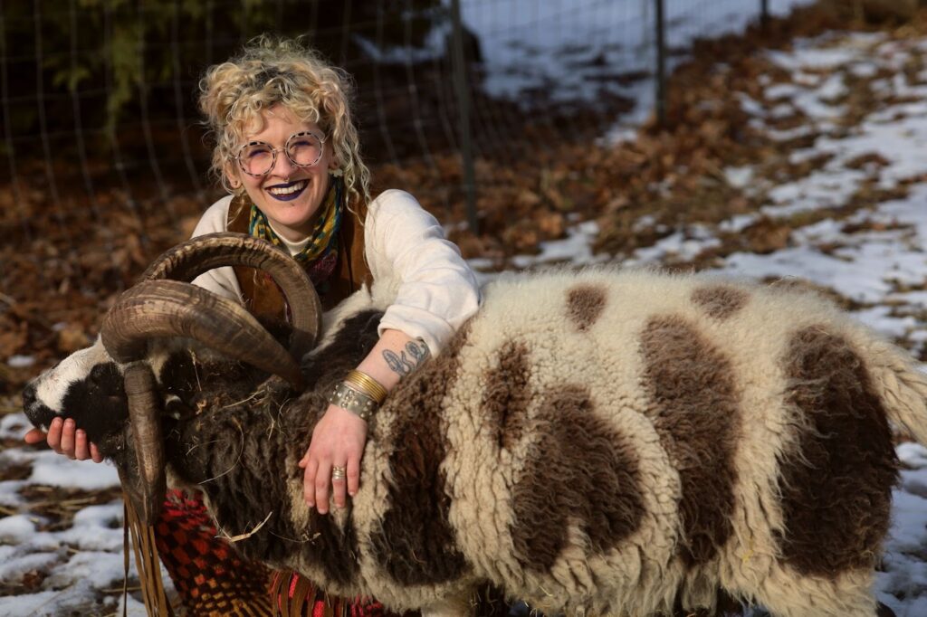Rachel kneels and smiles in the snowy pasture with Charlie, their four-horn Jacob ram. Rachel’s left arm is draped around his upper body as her right hand holds his chin. Charlie’s wool coat is speckled with large chocolatey brown spots and is peppered with a few pieces of loose hay. Image by Viki Stark.
