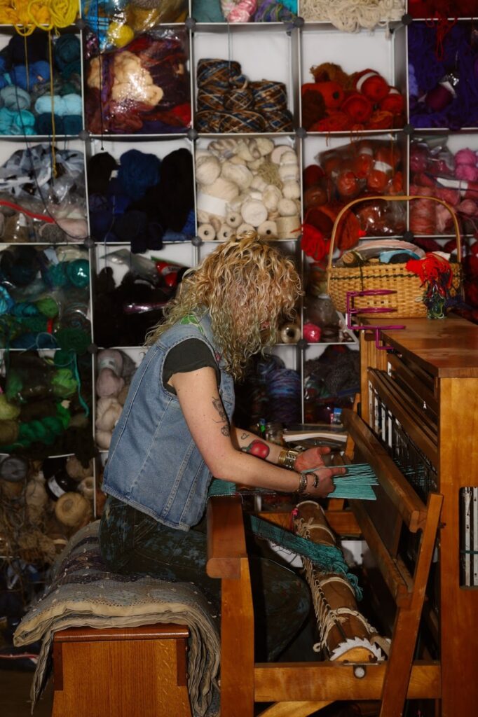 Rachel sits on a quilted bench as they weave a Double Two Tie Sampler on their 16 Shaft Leclerc Loom, which they had driven to New Jersey to pick up from a weaver who hand-wove samples for industry production. A packed shelving wall is seen behind her, fully stocked with hundreds of skeins of yarn, arranged by color. Image by Viki Stark.