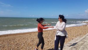 Shivani and her mother, Sanyukta, candidly photographed on a rocky beach. Wind blows their dark hair loose from their ponytails and creates whitecaps on the waves that crash towards shore. Shivani's arms reach out toward her mother, whose arms are crossed over her chest. Image courtesy of Shivani Gupta.