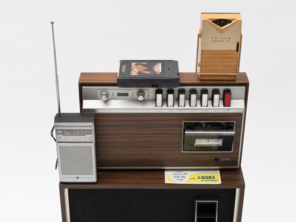 A detail shot of Jen Everett's piece Untitled, from the series Unheard Sounds, Come Through, 2025. Found objects. Dimensions Variable. Installation: 30 x 16.25 x 15". The image shows a stack of vintage stereos and other vintage audio equipment. Courtesy of the artist and Rivalry Projects, Buffalo, NY.