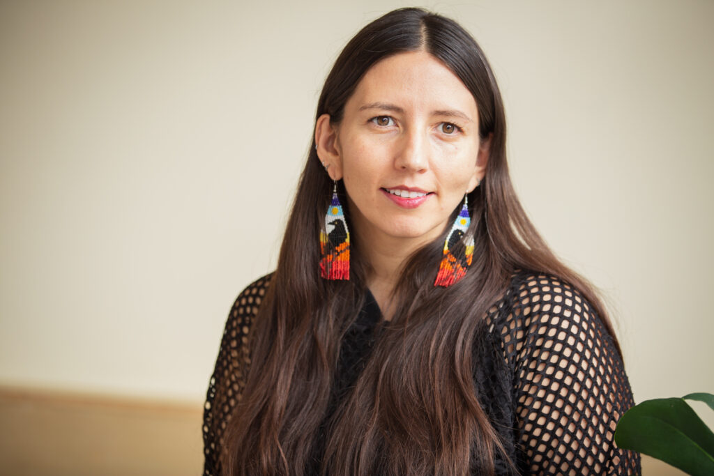 Crystal Vance Guerra sits at Tala Gallery while looking directly at the camera with a smile. She is wearing a black netted long sleeve shirt with colorful beaded earrings of crows perched on a branch. Image by Kristie Kahns.