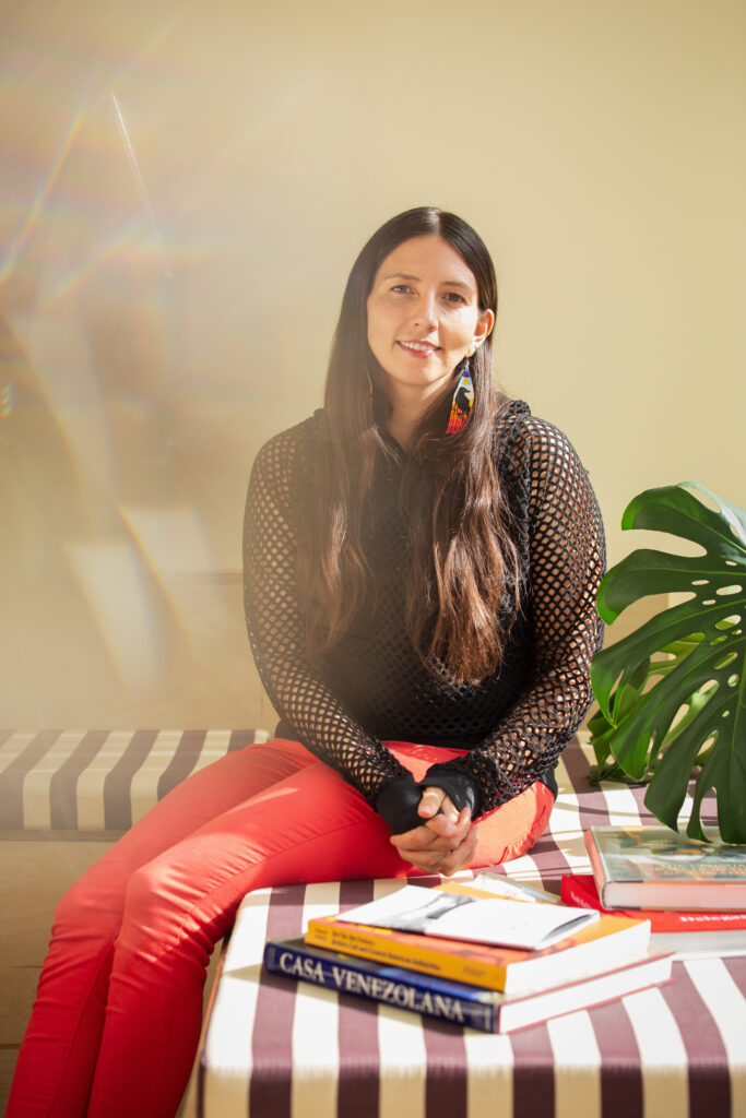 Crystal Vance Guerra sits on a bench in the front room at Tala Gallery with a stack of books at her side. The centered book in this image is Casa Venezolana. Image by Kristie Kahns.