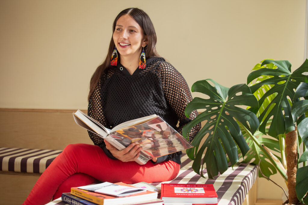 Crystal Vance Guerra sits on a bench in the front room at Tala Gallery with a large book in her hands and a scattered stack of books at her side. She is wearing a black netted long sleeve with red pants and is looking away from the camera with a smile on her face. Image by Kristie Kahns.