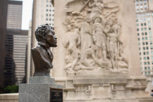 Image: A photograph of the profile of the DuSable bust created by sculptor Erik Blome in downtown Chicago. The bronzed bust sits atop a speckled stone pedestal with a black and gold plaque affixed below. Due to the angle of the photo, the words on the plaque are not easily legible. Blurred, behind the bust is a relief sculpture and skyscrapers. Photo by Kristie Kahns Photography.