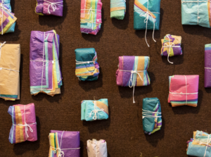 A board with tied up bundles on a wall. Layers and layers of colored tissue paper have been folded into bundles of different sizes, shapes, and colors. They are tied with a white string in various ways and pinned onto a textured, brown wall. Photo by Kristie Kahns.