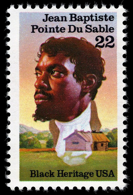 Image: The stamp featuring Dusable's likeness. Text at the top reads: "Jean Baptiste Pointe DuSable 22" DuSable's image is superimposed on an image of a simple cabin on green plains. The bottom text reads "Black Heritage USA." Image from The Smithsonian National Postal Museum.