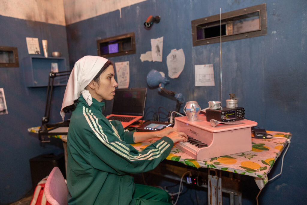 A portrait of Nadya Tolokonnikova for POLICE STATE at the MCA Chicago. A woman wearing a green Adidas tracksuit and wearing a white headdress plays a pink tabletop keyboard. Recording equipment, cords, and other instruments clutter the table as well. She is seated in a room resembling a prison cell. There are several drawings tacked to the blue wall. Image by Alexis Ellers.