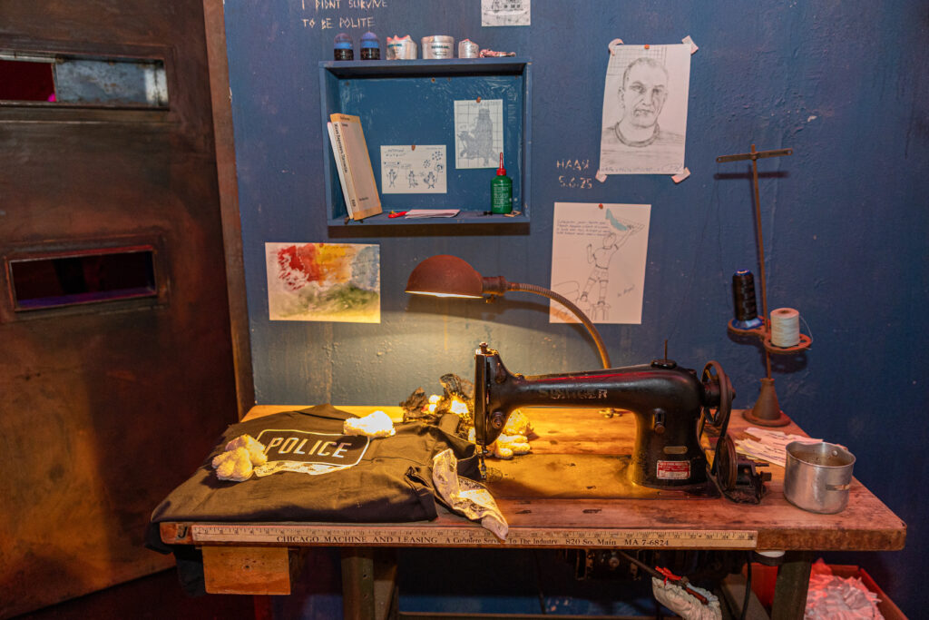 An installation view of POLICE STATE by Nadya Tolokonnikova. A ruler is attached to the side of a sewing table. A police uniform shirt with teddy bears sewn onto it is folded on the left side of the table. There is a lamp casting a yellow glow and a mug on the table. There are several drawings affixed to the wall, as well as an engraving in the wall that reads "I didn't survive to be polite." Image by Alexis Ellers.