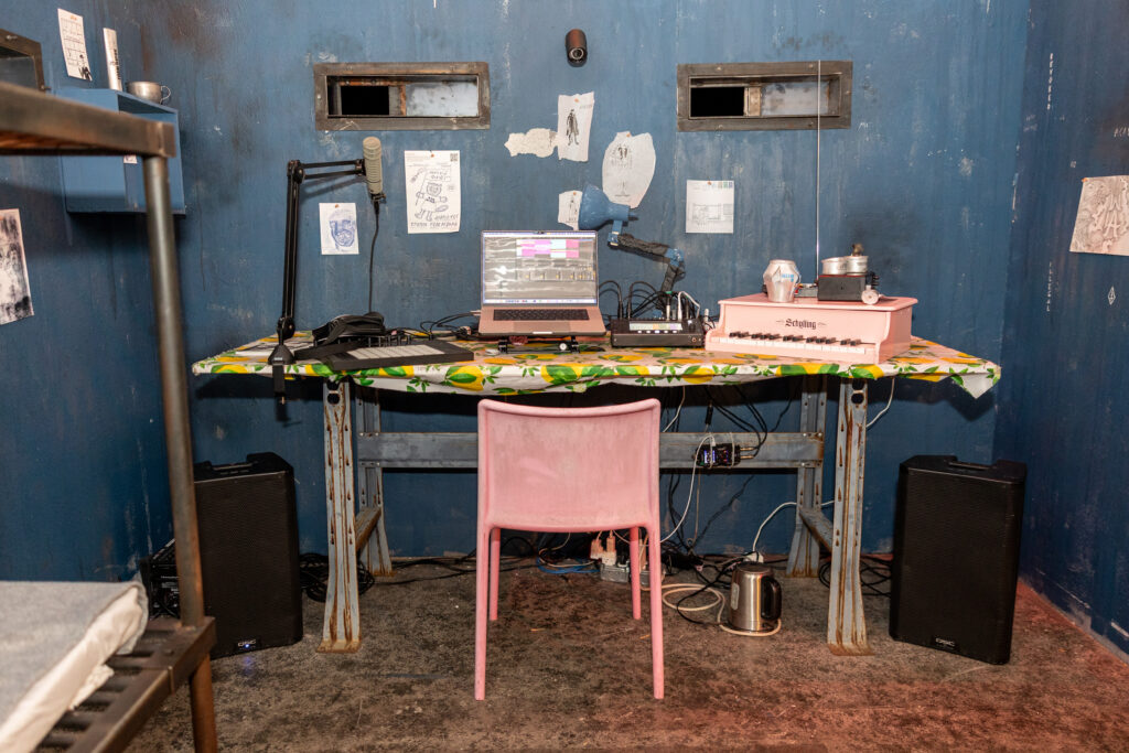 An installation view of POLICE STATE by Nadya Tolokonnikova at MCA Chicago. A makeshift recording studio is assembled in a room resembling a prison cell. Two speakers flank a metal workbench with a laptop, microphone, keyboard, and synths. There is an electric kettle on the gray carpet. There is a tablecloth with yellow lemons, a pink plastic chair, and several drawings pinned to the wall. Image by Alexis Ellers.