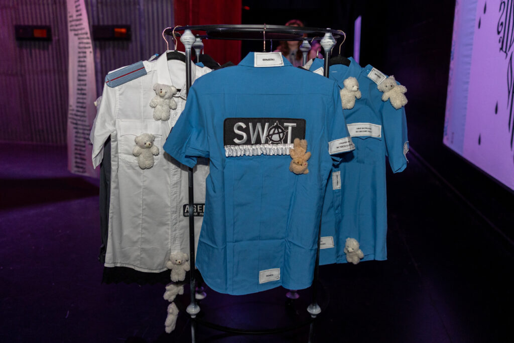 An installation view of POLICE STATE by Nadya Tolokonnikova at MCA Chicago. Several unironed prison guard shirts hang from a round display. One of the blue shirts has a patch that says "SWAT" with a circle-A symbol. This shirt, as well as a blue shirt and white shirt on either side, has small teddy bears sewn onto it. Image by Alexis Ellers.