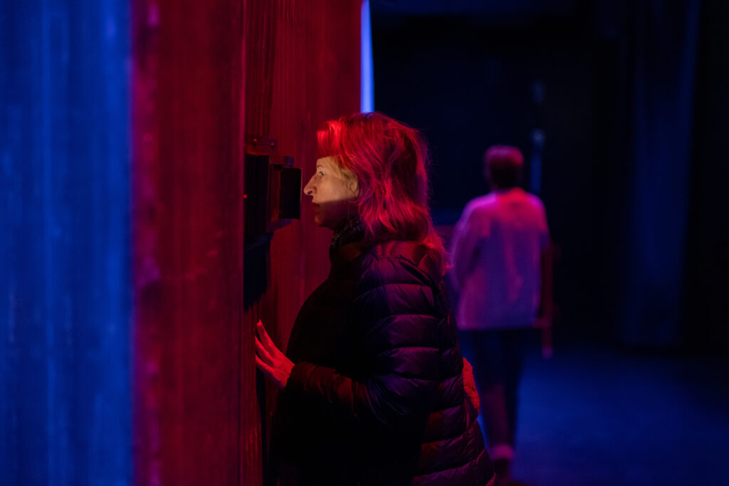 Image: An installation image of POLICE STATE by Nadya Tolokonnikova at the MCA Chicago. A woman wearing a black puffer jacket peers through a viewing port in the side of a sheet metal structure. Her face is illuminated by light from within the structure. A blurry figure can be seen in the background. Image by Alexis Ellers.