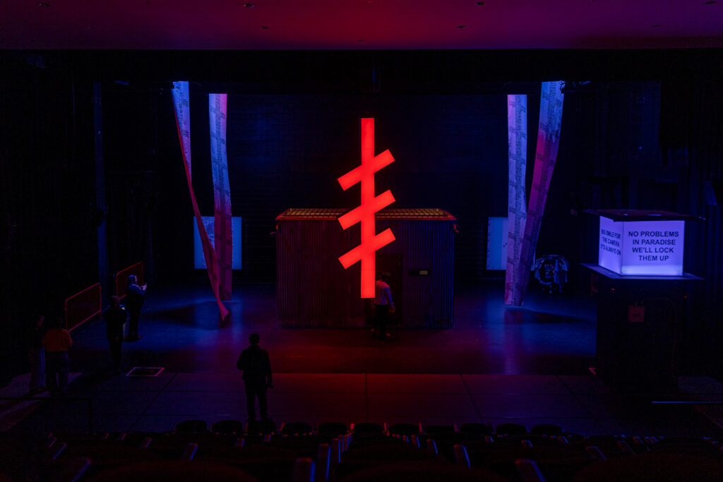 An installation view of POLICE STATE at MCA Chicago. A large, red neon sign interpreting an Orthodox Christian symbol hangs downstage in front of a corrugated metal structure flanked by long, hanging scrolls on either side of the structure. A sign on the right side of the stage reads, "NO PROBLEMS IN PARADISE WE'LL LOCK THEM UP." Half a dozen audience members are inspecting the metal structure.