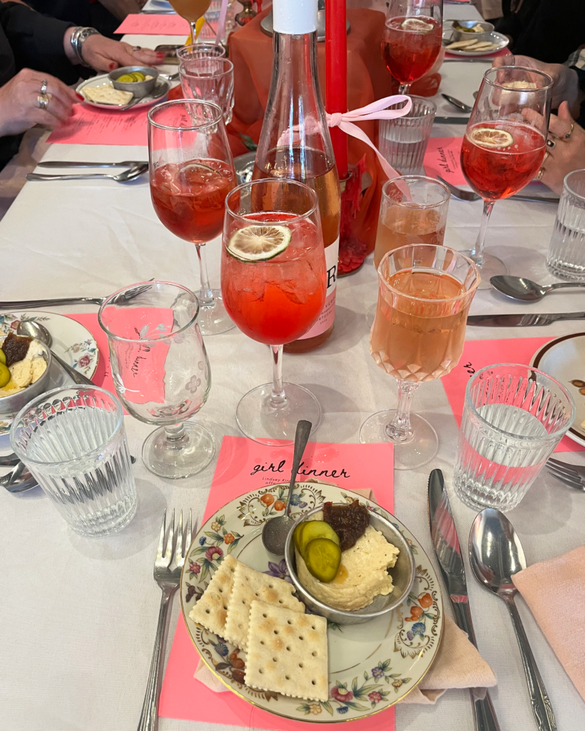 Image (right): A diner's view of the first course at Girl Dinner (white cheddar, nam prik pao, lime pickles, and saltines). Red-colored drinks in a variety of glasses and pink menus cover the table. Image courtesy of Lindsey Kircher.