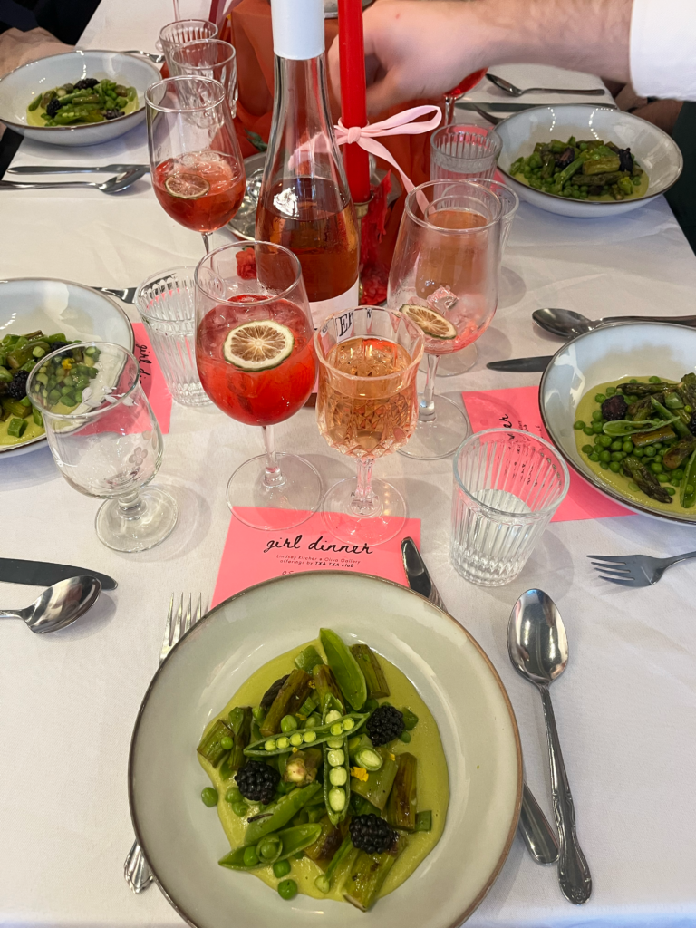 Image: A diner's view of the second course at Girl Dinner (spring peas, asparagus, blackberries, ramps, and egg). Red-colored drinks in glasses and pink menus can be seen nearby. Image courtesy of Lindsey Kircher.