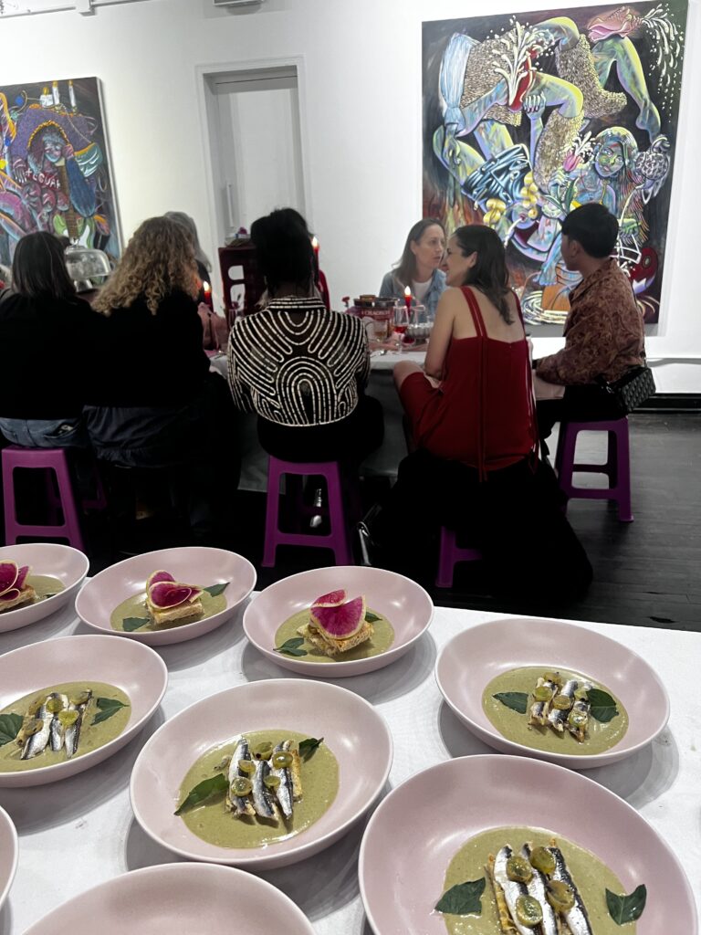 Image (left): A view of the final savory dish of the night at Girl Dinner (Spanish White Anchovies on toast in a green curry with grilled grapes, hibiscus, and Thai basil). A group of people sitting around a dinner table and two large paintings by Lindsey Kircher are hung on the walls behind them. Image courtesy of TXA TXA Club. 