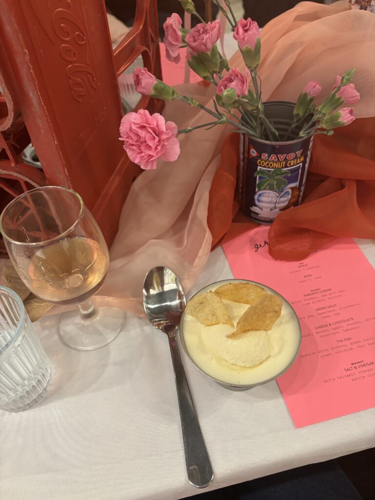 Image (right): A diner's view of desert at Girl Dinner (white balsamic vinegar ice cream served with kettle chips on top). Glassware, cutlery, flowers arranged in a coconut cream tin can, and reddish-orange chiffon cloth can be seen nearby. Image courtesy of Ally Fouts.