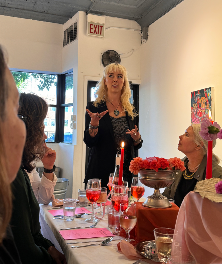 Image: Artist Lindsey Kircher stands while talking about her work to the attendees at Girl Dinner. She stands at the head of a table decorated with flowers, burnt-orange chiffon cloth, red candles, pink menus, and more. The dinner guests look at her. Image courtesy of Lindsey Kircher.