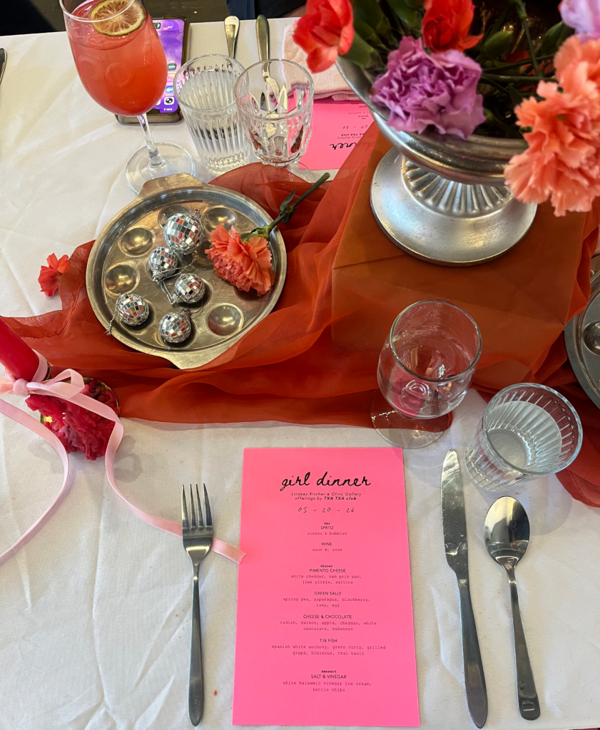 Image (left): A view of the tabletop at Girl Dinner looking down upon the pink menu. Cutlery, glassware, flowers, a orange-red scarf, and a silver tray of mini-disco balls can be seen across a white table cloth. Image courtesy of Lindsey Kircher. 