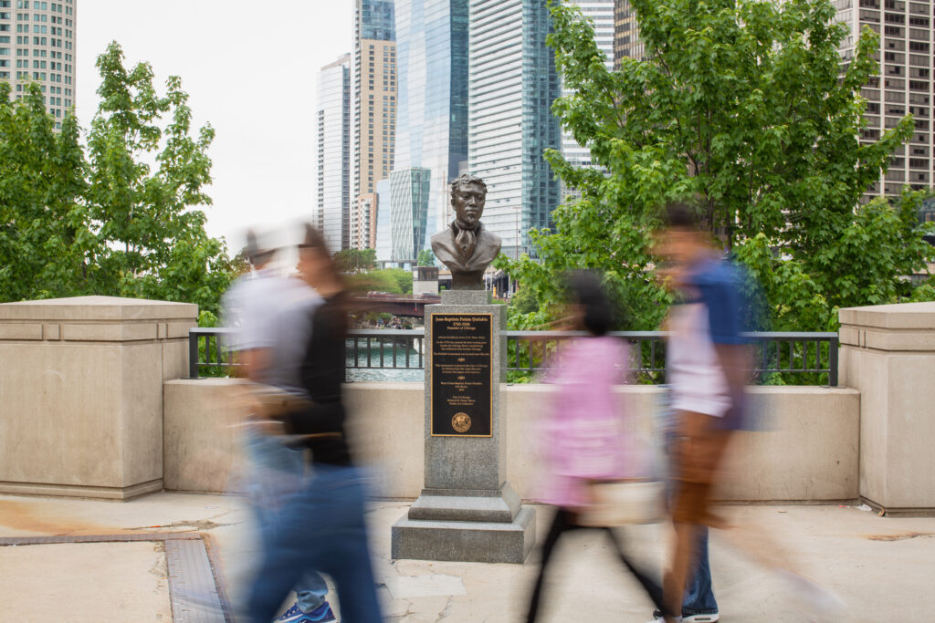 Image: A wide shot photograph of the DuSable sculpture by Erik Blome on the DuSable Bridge in Chicago, IL. Blurred images of pedestrians walk past the statue. Trees, the  riverfront, and skyscrapers can be seen in the background. Photo by Kristie Kahns Photography.