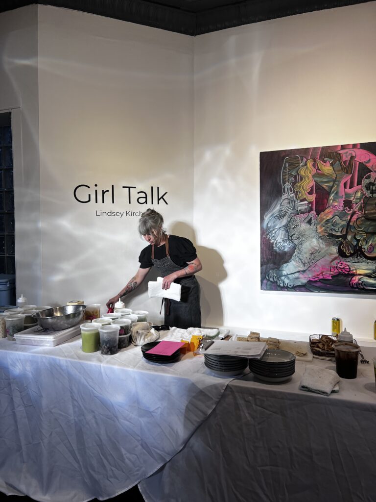 Image (left): A view of a food prep table at Girl Dinner. The words "Girl Talk" can be seen on the wall behind a person wearing an apron. A large painting by Lindsey Kircher can be seen on the right. Image courtesy of TXA TXA Club.