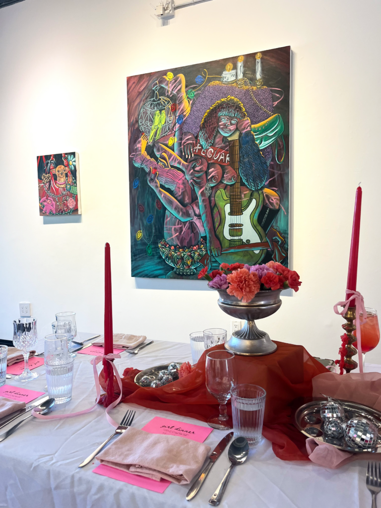 Image (right): A view of the tablescape at Girl Dinner, which included cutlery, pale pink napkins set on top of bright pink menus, tapered red candles, glassware, flowers, a orange-red chiffon cloth, a silver tray of mini disco balls, and more. Two paintings by Lindsey Kircher are hung on the wall in the background: Grotto Girl and Lap Dog. Image courtesy of Lindsey Kircher.