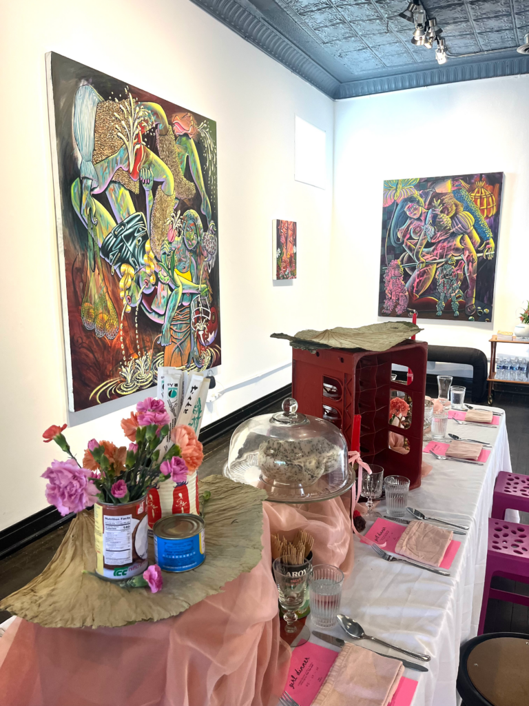 Image (left): A view of the tablescape at Girl Dinner, which includes four place settings and objects like a draped pale pink chiffon cloth, a cake stand, flowers arranged in tin cans, pink menus, cutlery, and more. Three paintings by Lindsey Kircher can be seen on view in the background. Image courtesy of Lindsey Kircher. 