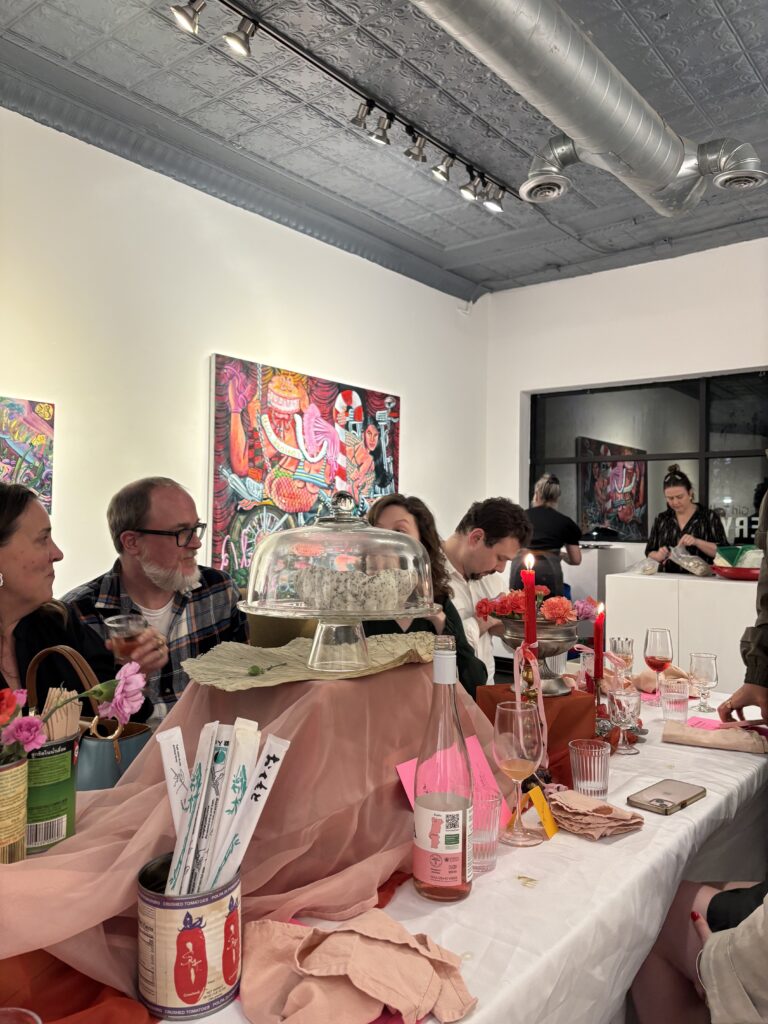 Image (left): A view of the dinner table at Girl Dinner with a group of people sitting and chatting. On the table are objects such as glassware, wine bottles, red candles, a cake stand, mauve-colored chiffon cloth, and more. Image courtesy of Ally Fouts.
