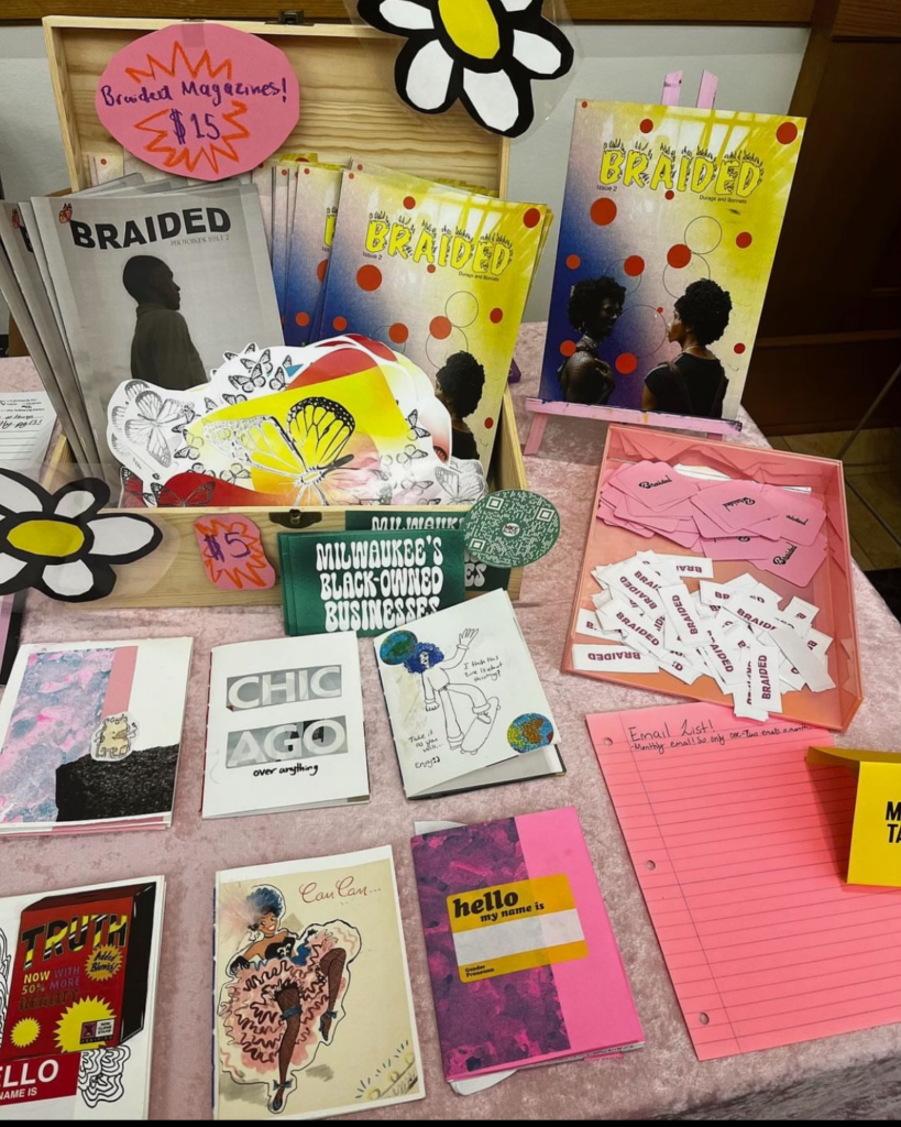 A photo of Ladasia Bryant's vending table at Milwaukee Zinefest. The top of the photo features Braided Magazine copies for sale for $15 and under it are $5 postcards and other miscellaneous zines for sale with an email list sign up sheet on the corner of the table. Photo courtesy of the author.