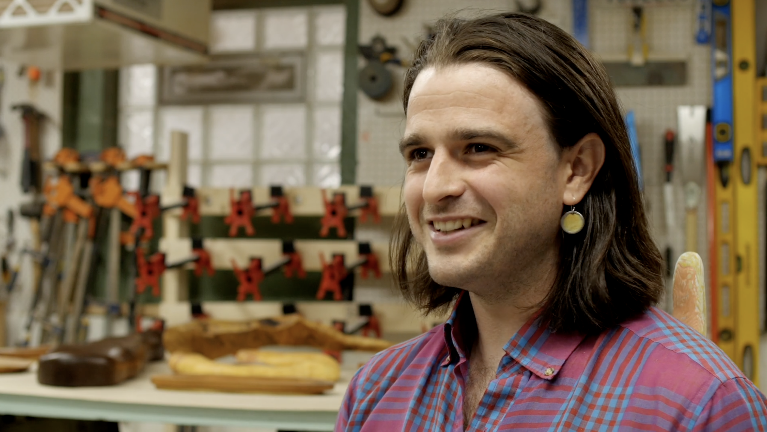 Forrest Hudes is seated facing the viewer's left and smiling. They wear a red and blue plaid shirt and have shoulder-length straight brown hair. Behind Forrest, a softly lit studio full of woodworking tools can be seen. Still image from a video interview by Ian Carstens.
