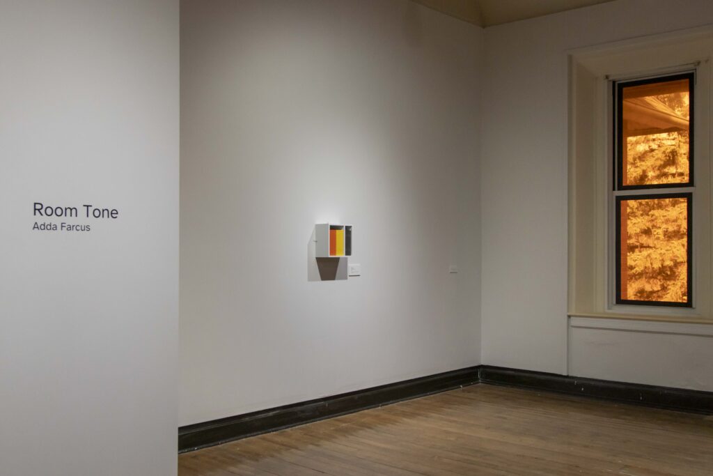 Installation view of Room Tone, Eureka College, 2026. On the left side of the image is the title of the exhibition with the artist's name underneath: 'Room Tone Adda Farcus.' Centered on a second gallery wall is a small, white, square shelf displaying zines with orange, yellow, and black bindings. They are grouped by color; orange on the left, yellow in the middle, and black on the right. Image courtesy of Adda Farcus. Photograph by Jade Minh-ha Nguyen.