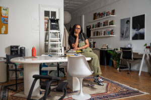 Image: OG Ugwuanyi clad in olive green overalls and a black t shirt sitting a top a white desk. The large space is filled with artworks and books. Photo by Tonal Mondae.
