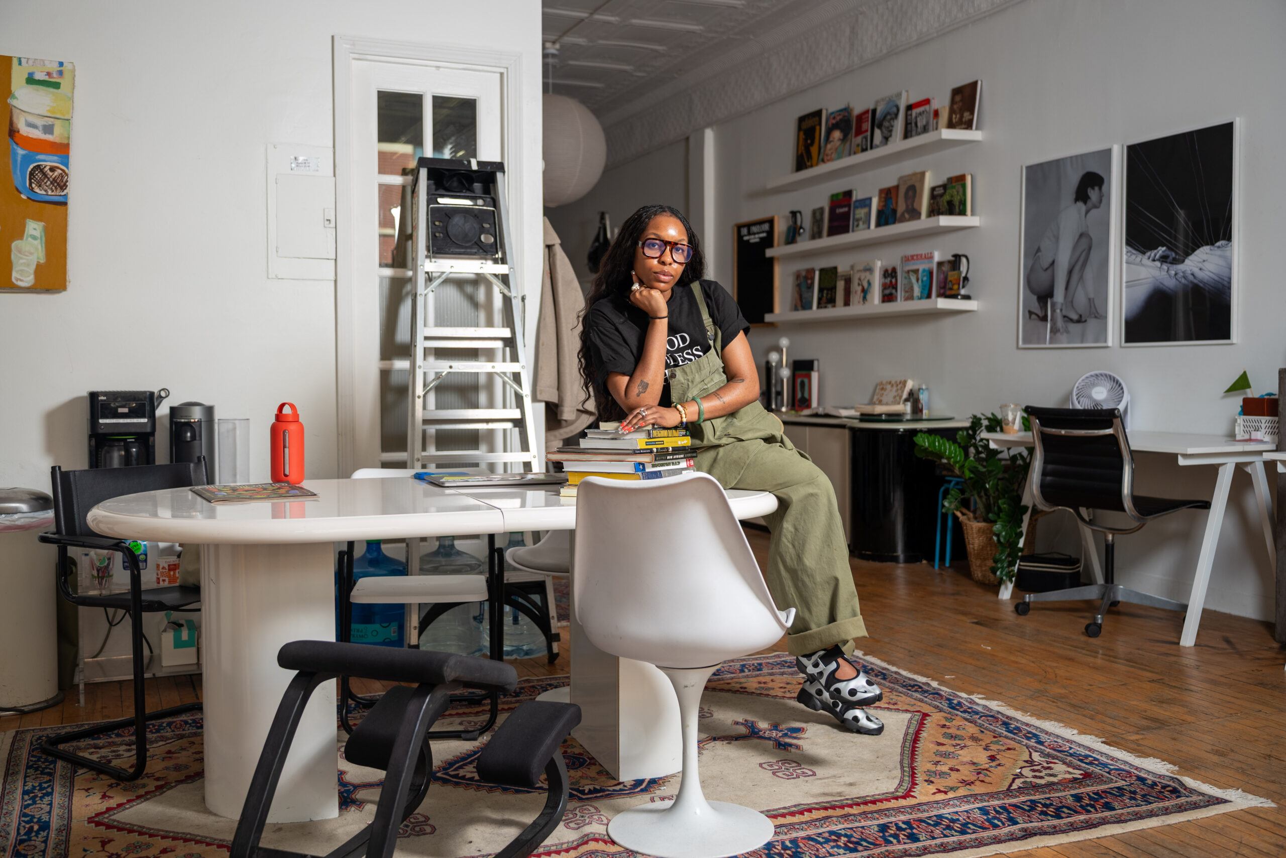 Image: OG Ugwuanyi clad in olive green overalls and a black t shirt sitting a top a white desk. The large space is filled with artworks and books. Photo by Tonal Mondae.