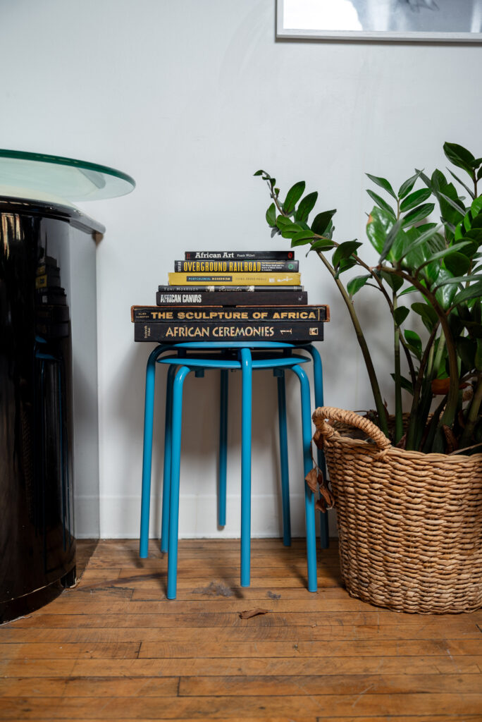 Seven books of various sizes and colors are stacked on top of a blue side table next to a wicker basket that contains a large potted plant. Photo by Tonal Mondae.