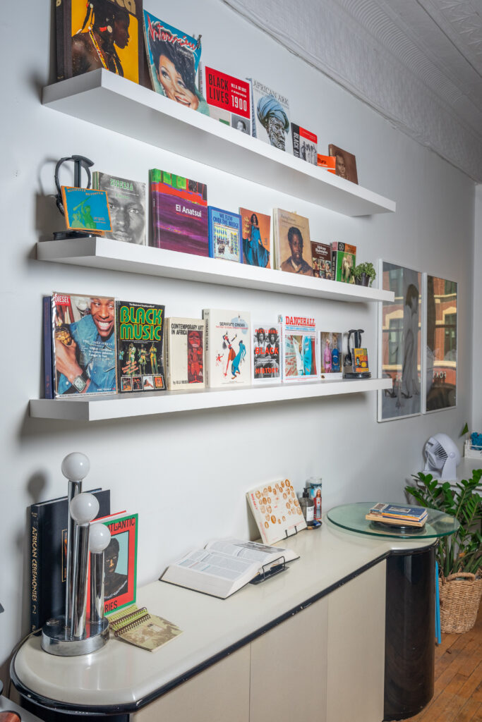 Three white floating shelves display an array of multicolored books and magazines above a midcentury modern credenza. Additional stacks of books are on the credenza, along with artwork and a lamp. Photo by Tonal Mondae.