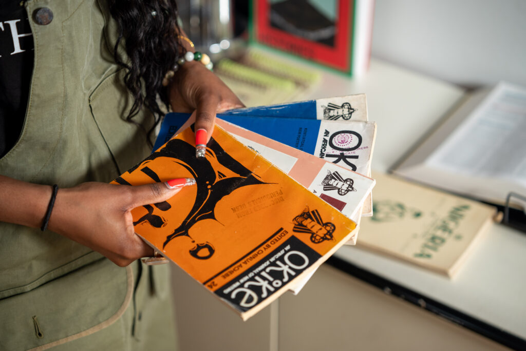 Close-up of OG holding four lightly weathered multicolored Okie booklets. OG's orange manicured nails complement the orange and blue book covers. Photo by Tonal Mondae.
