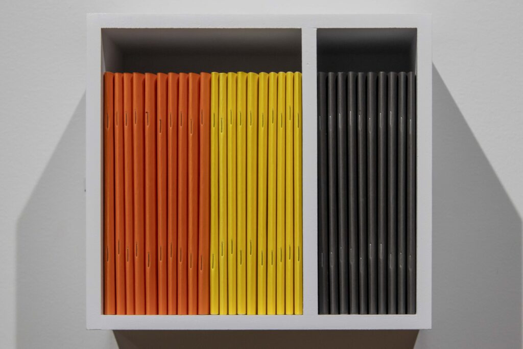 Adda Farcus, Wildfires, vol. 1, Wildfires, vol. 2, Psychoterratic Poems, zines, shelf, 2025-2026. A close up of the small, white, square shelf displaying zines with orange, yellow, and black bindings facing the viewer. They are grouped by color; 10 orange on the left, 10 yellow in the middle, and 10 black on the right. The yellow and black zines are separated by a white barrier that is part of the shelf. Image courtesy of Adda Farcus. Photograph by Jade Minh-ha Nguyen.