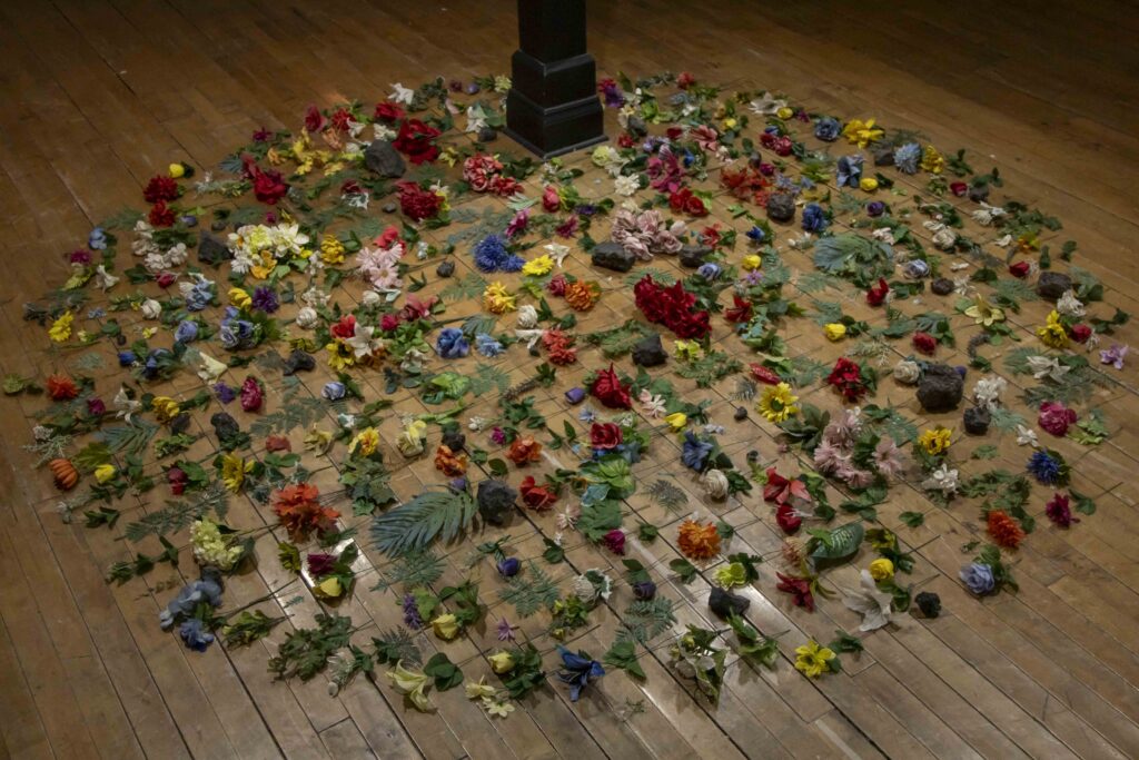 Adda Farcus, Wake, discarded cemetery flowers, clinkers, 2024. On an old hardwood floor are colorful flowers with green leaves, black chunks of rock-like fragments, and other found objects displayed in a large circle. The stems of the flowers are all pointed in the same direction. Image courtesy of Adda Farcus. Photograph by Jade Minh-ha Nguyen.