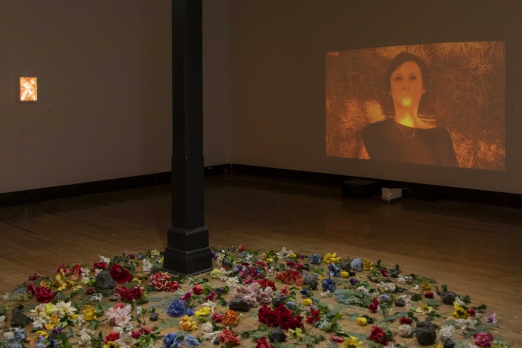 Image: Adda Farcus, death doula Ames Meeks chose this color to represent their feelings about climate change, video, 6:31, 2026. A still of Ames Meeks is projected onto a gallery wall in the color they chose that represented their feelings about climate change, 512 - Amber Delight. On a gallery wall to the left is another orange-colored piece from the exhibition. Below the video projection on the floor are some of the the colorful flowers with green leaves, dark chunks of various sized rock-like fragments, and other found objects from the piece Wake. Image courtesy of Adda Farcus. Photograph by Jade Minh-ha Nguyen.